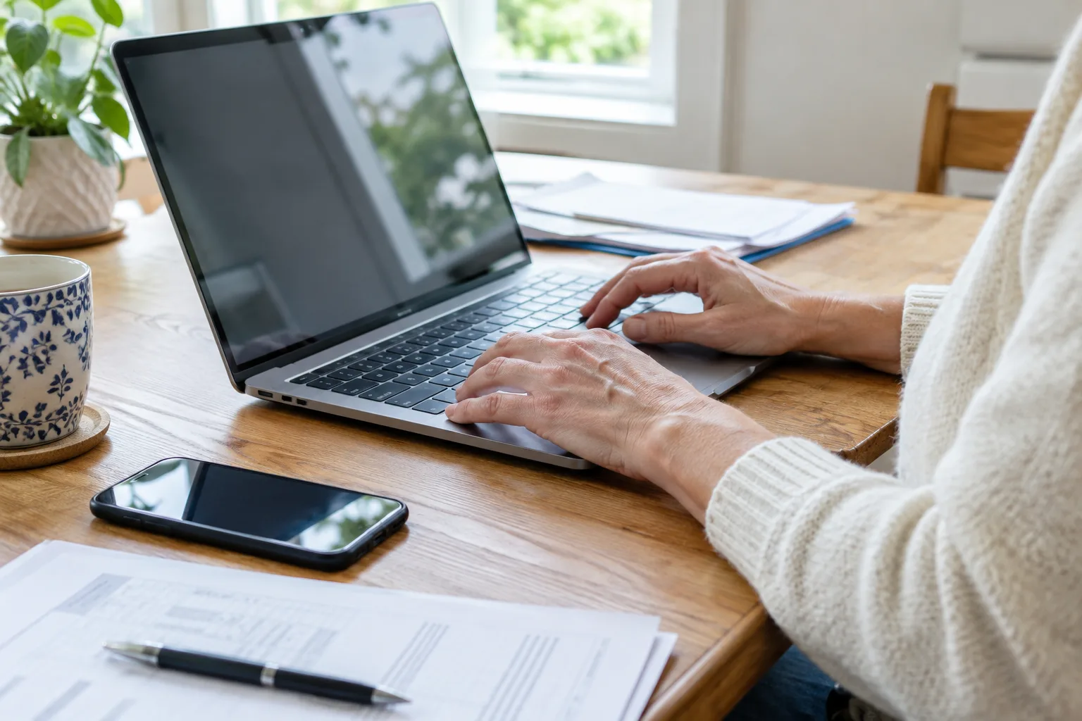 Des mains utilisent un ordinateur portable pour accéder à une démarche en ligne, avec un téléphone et des documents à proximité.